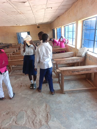 The image depicts an interior view of a classroom in Mada, Nigeria. The room features light-colored, worn walls with visible writing and graffiti, including "HON. HENRI ADAGAI" and "SENATOR 2007" on the left wall, and "CLAP JINEN ACHIEVE/CDC/O" on wooden desks. The ceiling consists of white, possibly water-stained, rectangular panels. The concrete floor is uneven, showing significant wear with exposed aggregate and rough patches.

Multiple wooden desks with integrated benches are arranged in rows, showing signs of use. Three rectangular windows with blue frames or grilles line the right wall, admitting bright natural light into the room. A light-colored backpack rests on the windowsill near the center right. A light blue doorframe is visible in the background on the left.

Seven individuals, possibly students, are present in the room. In the foreground, partially visible on the far left, a person wears white trousers and a pink backpack. Centrally, a female individual with dark skin wears a white long-sleeved shirt, a dark blue pleated skirt, white socks, and dark sandals. She has a white head covering and holds an open book or pamphlet in front of her face, partially obscuring it. To her right, a male individual with dark skin wears a white long-sleeved shirt and blue trousers, with dark sandals, standing with his back to the camera. Another male individual is partially visible behind him. Towards the right side of the room, near the windows, three individuals are dressed in bright pink long-sleeved garments. One is bending over a desk, another stands near a window, and the third is partially obscured further right. The individuals appear engaged in various activities within the classroom setting.