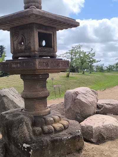 The image depicts a weathered stone lantern in what appears to be a park or garden setting. The lantern is a multi-tiered structure, with the main body being a square box with openings on each side, featuring decorative carvings. It rests on a decorative pedestal, which in turn sits on a rough-hewn stone base. Several large, rough-textured boulders are scattered around the base of the lantern. The background consists of a gently sloping grassy hill dotted with trees, and a pathway winding through it. The sky is partly cloudy with patches of blue. The overall scene is naturalistic and serene. The location is identified as Mažučiai, Lithuania.
