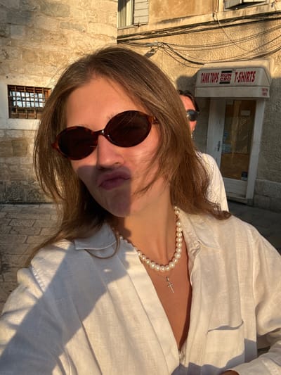 This is a vertical, close-up, waist-up photograph of a young woman taken outdoors in Split, Croatia, during what appears to be the golden hour, given the warm, low-angled sunlight casting long shadows. She is wearing a white linen shirt and a pearl necklace with a small silver cross pendant. Her hair is brown and falls around her shoulders. She has on large, dark, tortoiseshell sunglasses that obscure most of her face, but her lips, painted a mauve shade, are visible.
The background suggests an urban setting with a stone building and a window with a barred grate. Above the woman's head, a white sign with red lettering is visible, featuring the text "TANK TOPS" and "T-SHIRTS", along with "RUN CRO". The overall mood is casual and relaxed, possibly capturing a moment of leisure or sightseeing in the historic Mediterranean city. The sunlight creates a soft, warm glow on the woman and the textures of her clothing and surroundings.