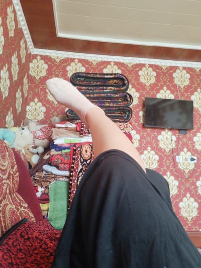 The image depicts an indoor scene in what appears to be a living room. A person's leg and foot, clad in a light-colored sock, are prominently featured, extending from the bottom right corner of the frame. The person is wearing a dark skirt. The setting includes a wall covered in red and gold patterned wallpaper, with a white decorative border. A small black television is mounted on the wall, with electrical outlets and wires visible below it. To the left of the television, a stack of folded blankets or textiles with intricate designs is visible. Further to the left, a pile of stuffed animals, including a white teddy bear, and more folded textiles are present, partially obscuring a red patterned sofa. The lighting suggests it is daytime. The location context provided is "Socialism, Kyrgyzstan," which may imply a style of decor or furnishings consistent with that era or region. No specific activity or event is clearly depicted beyond the static arrangement of objects and the person's leg. There is no visible text in the image.