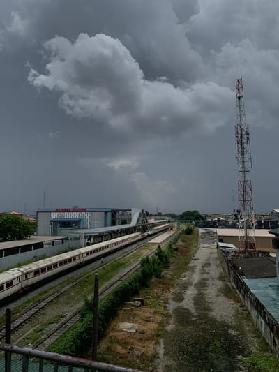 The image depicts a railway scene in Ebute-Metta, Nigeria, under a cloudy sky. A tall, red and white lattice tower stands in the distance. Alongside the railway tracks, there is a station building with the name "OYINGBO STATION" visible. A white and red train sits on one of the tracks. There is some green vegetation adjacent to the tracks and a concrete wall along the right side of the image.
