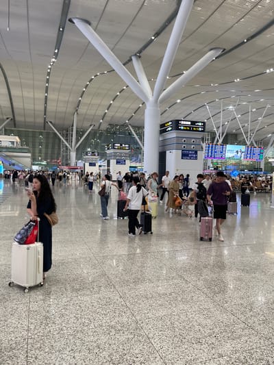 This media file captures a bustling scene inside a modern airport or train station, likely in China's Longhua District given the provided context. The wide-angle shot reveals a long, expansive hall with a high, arched ceiling supported by large, white, futuristic-looking pillars. The floor is a speckled granite, reflecting the ambient light.
Numerous people are present, some standing, some sitting, and others walking with luggage. They are diverse in age and attire, suggesting a mix of travelers. Many are seen with rolling suitcases, indicating they are either waiting for or have just arrived from a journey. The overall atmosphere appears busy but orderly, with people queuing or relaxing while waiting for their departures or arrivals.
Visible text in the image includes directional signs and electronic display boards. One sign reads "6-20 68-78 | 6A-7A" and another indicates "←B6-B7 A1西 认养1头牛". These signs likely provide gate numbers, platform information, and possibly advertising. The presence of these signs and the large waiting area strongly suggest a transportation hub.
The lighting appears to be artificial and diffused, creating a bright, indoor environment. The time of day cannot be precisely determined but the activity level implies it's during operational hours. No specific emotional expressions are clearly discernible from the crowd, but the general mood seems to be one of anticipation or patient waiting. A woman in the foreground is looking at her phone, a common sight in such settings. The overall impression is that of a well-organized and active travel terminal.