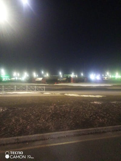At night in Egor'evo, Russia, a partially snow-covered landscape is illuminated by distant streetlights. A low white fence separates the foreground from the illuminated area. The foreground features a dark, mottled patch of ground bordering a paved road. In the background, illuminated structures and signs are visible beyond the fence, along with what appears to be construction materials or large pipes. A gas station is visible on the left.
