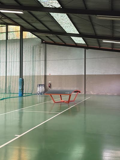 This image displays an unoccupied indoor sports facility located in Santarém, Portugal. The scene features a green, glossy sports court marked with white lines, reflecting overhead light. Centrally positioned in the midground is a dark gray teqball table with an orange-red, curved base, bearing the "TEQ" and "Q" logos. To the left, a tall green mesh net spans vertically, secured by a dark pole padded in blue at its base. Behind the net, against the wall, a blue metal rack containing white bags is visible, alongside a red fire extinguisher mounted on the two-toned wall. The walls consist of a light gray upper section and a darker, textured lower section. The ceiling features a corrugated metal roof structure supported by dark beams, incorporating several rectangular skylights and linear fluorescent light fixtures. No individuals, actions, or interactions are present in the frame.