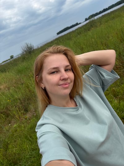 A young woman with blonde hair and freckles is taking a selfie outdoors. She is wearing a light blue t-shirt and has her arm behind her head. She is smiling gently at the camera. Behind her, there is a vast expanse of green grass leading to a body of water under a cloudy sky. The overall scene suggests a natural, possibly rural, setting. Given the context of Breytovo, Russia, this could be a scene by the Rybinsk Reservoir.