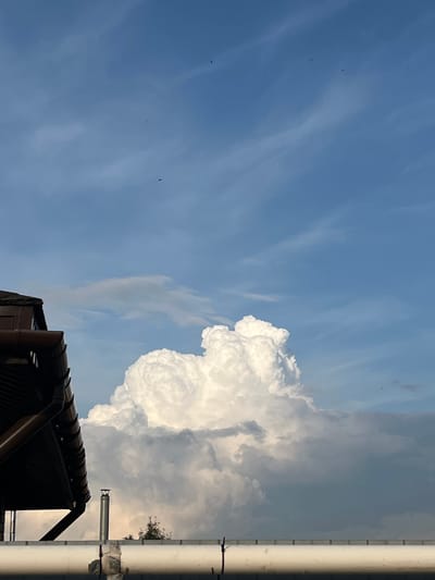 The image depicts a view of the sky with large, white cumulus clouds dominating the center. In the foreground on the left, a portion of a dark-brown roof with gutters is visible. Below the roofline, a grey pipe or conduit runs horizontally across the lower part of the frame. A metallic chimney with a cap is situated on the right side of the frame, slightly behind the horizontal pipe. A few dark specks, likely birds, are present in the blue sky. The sky is partly cloudy with streaks of lighter clouds. The scene appears to be an outdoor setting, possibly a rooftop or elevated vantage point, in an urban or suburban environment given the presence of building structures and infrastructure. The lighting suggests it is daytime, with the sun illuminating the clouds from the side. The location is identified as Сухая Балка, Russia.