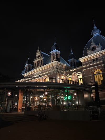 This nighttime image depicts the Musis Sacrum complex in Arnhem, Netherlands. The primary subject is a multi-story, historic building, the Musis Sacrum concert hall, constructed of reddish-brown brick with cream-colored architectural trim. It features multiple dark, domed roofs and turrets, each topped with a decorative finial. Several arched and rectangular windows are present, emitting warm yellow light. A prominent inscription reading "MUSIS SACRUM" is visible on an upper facade section. An elevated, curvilinear walkway with an ornate metal railing extends along the building's mid-section.
Below this structure, a modern, glass-fronted cafe or restaurant is situated. Its interior, featuring tables and chairs, is brightly lit with warm-toned illumination. Several distinct, pendant-style light fixtures with glowing globes hang outside the cafe's glass walls. A green neon sign spelling "JANS" is visible within the cafe. A horizontal band above the glass facade displays repeating text, including "CAFÉ RESTAURANT FOYER TERRAS."
On the paved ground in front of the cafe, a dark-colored bicycle is parked upright. An outdoor seating area with tables and chairs is arranged on the dark ground. A large, dark folded outdoor umbrella is positioned to the right of the cafe's outdoor area. The sky above is uniformly dark, indicating nighttime conditions. No individuals are present in the visible scene.