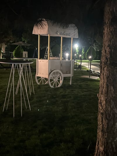 In Vyshen'ky, Ukraine, at night, a white cart with gold accents and wheels stands on a grassy lawn. The cart is adorned with a white awning that has "La Dolce Vita" written on it in script. A ladder leans against the cart. In the foreground, a round table with a black top is supported by thin white legs. In the background, trees and lights are visible.