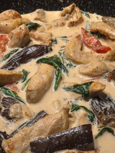 The image shows a close-up, overhead view of a dish being cooked in a dark pan. The dish appears to be a creamy stew with several ingredients including pieces of what looks like chicken, wilted spinach, slices of eggplant, and small red pieces that might be tomatoes. The creamy sauce has a pale yellow to off-white color and is frothy in places, indicating it is hot and simmering. The lighting suggests it is daytime, possibly indoors in a kitchen setting. There is no visible text in the image. The focus is entirely on the food, highlighting its texture and components. The dark, speckled surface of the pan is visible around the edges of the dish. The overall impression is of a rich, savory meal being prepared.