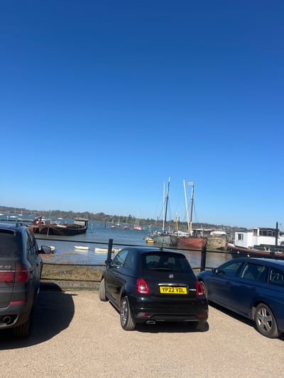 The scene shows a waterfront area in Babergh, United Kingdom, on a clear day with a cloudless blue sky. Three cars are parked along a waterfront street. A black Fiat 500 with license plate YP22 VDL is parked between two larger, darker cars. The Fiat is positioned directly next to a low fence that runs along the water's edge. Beyond the fence, a body of water (likely the River Stour) is visible with several boats moored. Some have masts. The edge of a building is visible on the far side of the water.