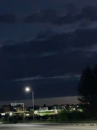 This is a nighttime image of a road with buildings and streetlights in the background. The sky is dark and cloudy, with a few illuminated clouds visible. A single, bright streetlight illuminates the foreground of the image, casting a glow on the road and surrounding vegetation. In the distance, several buildings are lit up, suggesting a residential or commercial area. The image appears to be taken from a slightly elevated position, possibly from a bridge or overpass, looking towards a town or city. There are no people or animals visible in the image. The overall atmosphere is calm and quiet, typical of a nocturnal urban landscape.