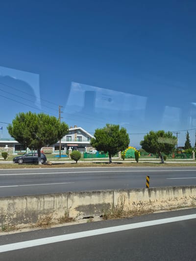 The image depicts a roadside view in Νέο Ρύσιο, Greece, on a clear day under a bright blue sky. In the foreground, a grey asphalt road runs horizontally, bordered by a weathered concrete barrier. A yellow and black striped traffic post stands on the barrier. Beyond the barrier, a strip of dry grass and weeds lines the edge of the road.

Further back, a paved area is visible, with a dark grey car parked on the left side. Behind the car, a building with a grey facade and a multi-pitched roof stands. The building has multiple windows and balconies. Trees with dense green foliage are planted in front of and to the side of the building. A playground with colorful structures, including a slide and swings, is situated to the right of the building. A metal fence encloses the property. Overhead power lines are strung across the scene. The overall impression is one of a suburban or semi-rural environment.