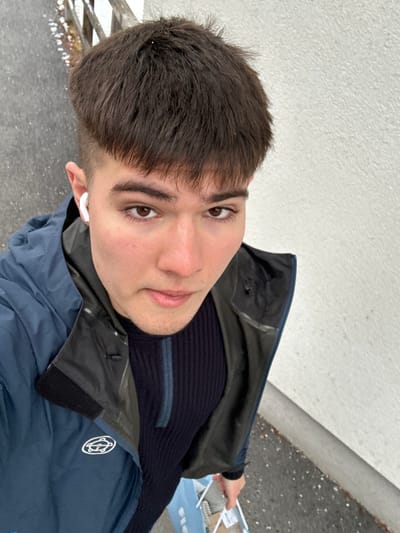 The image displays a head-and-shoulders portrait of a young man, rotated 90 degrees counter-clockwise. He has short, dark brown hair with a fringe covering his forehead, and brown eyes. He is looking directly at the camera with a neutral to slightly smiling expression. His left ear is visible, featuring a white earbud.
He is dressed in a dark blue or black ribbed knit sweater with a horizontal lighter blue or grey stripe across the chest. Over the sweater, he wears a multi-toned jacket; the main body is dark blue, while the upper chest and shoulder area appears to be a slightly different, possibly greenish-grey, material. A white, circular logo with an abstract, stylized pattern is visible on the left side of his chest on the jacket.
The background behind him consists of a light, possibly white or light grey, textured wall to his left and a darker grey ground surface to his right. A hand, presumably his, is visible on the far right of the image, holding a light blue bag that contains some light-colored items, possibly purchases or groceries. The lighting suggests an outdoor, daytime setting, possibly overcast, as there are no strong shadows.