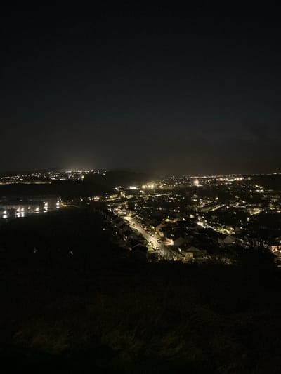 Night aerial view of Gourock, United Kingdom. City lights illuminate houses and streets along the coast. Dark sky above the city. Shadowy hill with vegetation on the right.
