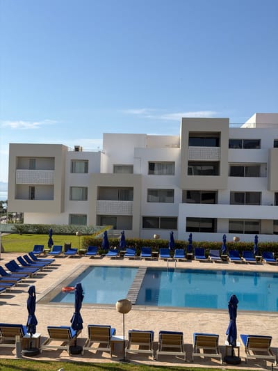 The image shows a swimming pool area in Nador, Morocco. A modern, white building stands adjacent to a rectangular swimming pool. The pool is lined with blue umbrellas and lounge chairs, and a grassy area is visible next to the pool deck. The sky is clear and blue. There are no people visible in the image. A life preserver is present in the pool.