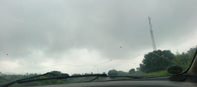 The image captures a view from inside a vehicle in Assob Hausa, Nigeria, looking out through a cracked and rain-speckled windshield on an overcast day. The car's dashboard and windshield wipers are visible in the foreground.
A prominent, large crack traverses the windshield horizontally across the middle and lower sections, with several finer cracks also apparent, particularly in the upper left. Numerous small water droplets or specks of dirt are scattered across the glass.
Beyond the windshield, the sky is a uniform, dull grey, indicative of heavy cloud cover. On the right side of the frame, a tall, red and white telecommunications tower rises above the tree line. The midground consists of a dense, green expanse of trees and foliage, suggesting a rural or natural landscape.
No people are visible. An oval-shaped green sticker, with text that is too indistinct to read, is attached to the dashboard area on the right, near the windshield frame. The scene conveys a moment of observation from a car, under gloomy weather conditions, with the damaged windshield being a central element of the visual narrative.