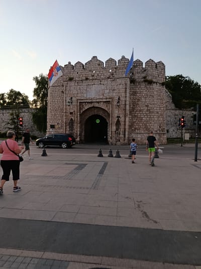 A Serbian flag and a blue flag fly from the top of a medieval stone gate. Two people, a man and a boy, walk away from the camera on a sidewalk in front of the gate. A black car is stopped at a crosswalk. A woman in a pink shirt and black shorts walks away from the camera on the sidewalk to the left of the gate. The sun is low in the sky, casting long shadows. The environment is an urban setting in Градска општина Црвени Крст, Serbia.