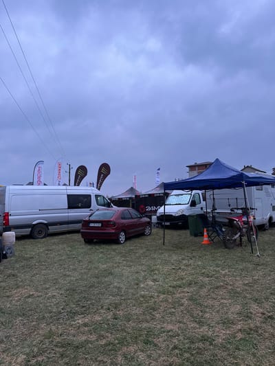 This image depicts an outdoor event or temporary setup within a grassy field in Poroy, Bulgaria, under an overcast sky.

The scene features multiple vehicles and temporary structures. On the left, a silver cargo van with dark windows is parked. Adjacent to it, a dark red sedan, bearing Bulgarian license plate H 0516 BA, is parked facing away from the viewer. Behind these vehicles, several tall, narrow advertising flags display logos including "BARDAH," "AVTOMOTO," and "MONSTER." Two black pop-up tents, branded with "24MX" in white, are also visible.

In the center-right, a white van with a sunshade in its windshield is parked. To its right, a blue pop-up canopy tent is erected, with a visible white recreational vehicle (RV) or motorhome parked behind it. Underneath or near the blue canopy, a red dirt bike is parked, accompanied by a folded camping chair and an orange traffic cone.

In the background, beyond the temporary structures, several residential buildings with tiled roofs are visible, indicating proximity to a developed area. No individuals are present or engaged in observable actions within the frame.