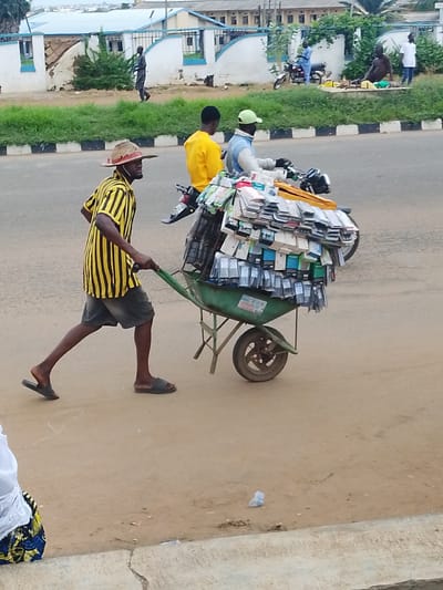 In Oja Oba, Oko Erin, Nigeria, a man wearing a straw hat and a yellow and black striped shirt pushes a wheelbarrow laden with what appear to be phone cases. He is wearing dark shorts and sandals and is walking on a dirt road. Behind him, another person wearing a yellow shirt is visible. In the background, there are buildings, a grassy embankment, and other people and motorcycles, suggesting a busy marketplace or street scene. The overall impression is one of commerce and daily life in a Nigerian town. The lighting suggests it is daytime, with no clear indication of the weather. No visible text is present in the image.