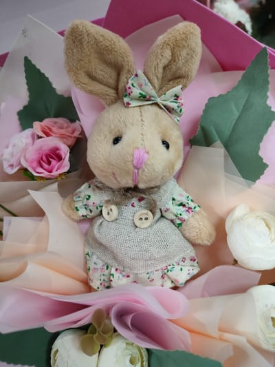 A plush bunny toy is presented in a bouquet, nestled amongst pink and peach-colored fabric roses and green leaves. The bunny wears a light-colored dress with a floral print and two wooden buttons. A pink bow with a floral pattern adorns its head. The overall impression is a gift arrangement, likely for a special occasion. The image does not contain any information that would allow for the determination of its location in the city of Kalach, Russia.