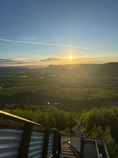 The image depicts a scenic landscape at sunrise, viewed from an elevated vantage point, likely a structure with corrugated metal siding visible in the foreground. The sun is low on the horizon, casting a warm golden glow across the sky and illuminating a vast expanse of dense green forest that stretches into the distance. Wispy clouds and a distinct contrail are visible in the clear blue sky.

The foreground shows the textured surface of corrugated metal and weathered wood, suggesting a man-made structure overlooking the natural scenery. The middle ground is dominated by the lush, rolling forest, with hints of clearings or bodies of water interspersed within the trees. In the far distance, a mountain range or large hills are silhouetted against the bright sky.

The time of day is clearly sunrise, characterized by the low angle of the sun and the soft, warm light. The weather appears to be clear with a few clouds. The location, given the context, is Oliver Paipoonge, Canada, a region known for its natural beauty and forested areas. There are no people or specific activities depicted. The overall mood evoked is one of tranquility and awe at the natural beauty of the landscape at the beginning of a new day. No visible text is present in the image.