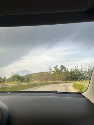 The image shows a view from inside a vehicle, likely a car, through a sunroof or window. Raindrops are visible on the glass surface. Outside the vehicle, there is a view of a road and a grassy hillside with trees. The sky is overcast with gray clouds. The photo was taken in Kipra, Bulgaria.