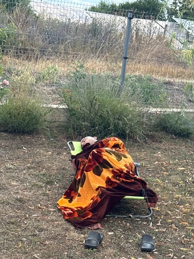 The image shows a person sleeping on a green chair outdoors, covered by a brown blanket with an orange and red floral pattern. Two black sandals are placed on the ground near the chair. The ground is covered in dry grass and fallen leaves. In the background, there is a wire fence and overgrown vegetation. The scene appears to be in a yard or garden area. The city of Vizitsa, Bulgaria, is not visually identifiable within the image.