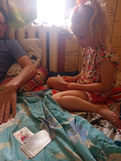 In a dimly lit room, likely in Gdov, Russia, a girl with blonde hair tied in a ponytail is engrossed in a game of cards. She wears a t-shirt with a strawberry pattern and red shorts. Her right hand holds a few cards, while her left hand is near a larger stack of cards. Across from her, a person's arm, resting on pillows, is visible, suggesting a shared activity. The environment appears to be a bedroom, with a window providing natural light, casting shadows on the wall. A radiator and a patterned blanket on the bed contribute to the domestic setting. The overall mood is one of quiet concentration and leisure.