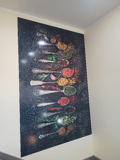 A rectangular artwork featuring a black, textured background adorns a wall. It displays an array of ornate silver spoons, each filled with a different spice or herb, like saffron, paprika, peppercorns, and rosemary. The spices create colorful accents against the dark backdrop. The arrangement is horizontally oriented.