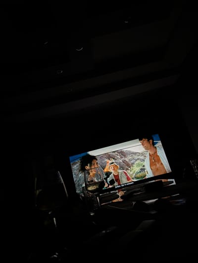 The image shows a dimly lit room, likely a home theater or living room, with a television screen displaying a scene from a movie or show. The screen shows two men, one wearing a hat and a red shirt under a light-colored jacket, and another man with an open white shirt, exposing his chest. The background on the screen appears to be a rocky, outdoor landscape with some greenery. There is text visible on the bottom of the screen which reads "Добре. Ще го направя." which translates to "Okay. I'll do it." In the foreground of the image, a glass with a clear liquid, possibly wine, is visible in silhouette. The room is dark, with some recessed ceiling lights faintly visible in the upper portion of the image. The overall impression is that of a person watching entertainment in a darkened environment, possibly alone or with others not clearly depicted. The location in Petarch, Bulgaria is not determinable from the image itself.