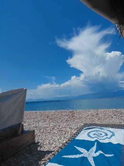 This is a horizontal, eye-level, outdoor shot of a beach in the Municipal Unit of Diakopto, Greece. The foreground is dominated by a pebbled beach, with a blue and white towel laid out on the right. The towel features a star-shaped design and a swirling pattern. To the left, a portion of a beach chair with a patterned cushion is visible. Beyond the beach is a calm, blue body of water, likely the sea, with a distant coastline of mountains under a bright blue sky. A large, white, puffy cloud formation is prominent in the sky. The image appears to be taken on a sunny day. There are no people visible in the frame.