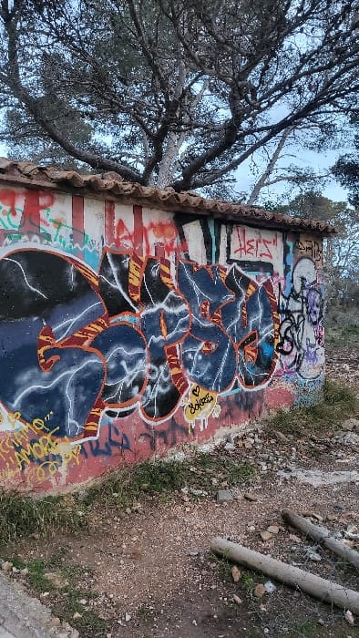 The image displays a graffiti-covered single-story utility building situated within an unkempt outdoor environment in Santa Ponça, Spain. The structure's walls are extensively tagged with multiple layers of spray paint, featuring a dominant piece in dark blue and black with white outlines and orange stripe accents. A yellow tag reading "Soket" with a heart symbol is visible beneath this main graffiti. The building is topped with a roof composed of reddish-brown clay tiles. Overhead, mature trees with dark, gnarled branches and sparse foliage rise against an overcast, light blue-grey sky. The ground in the foreground is uneven, consisting of reddish-brown soil, gravel, scattered stones, and patches of dry green grass. Two weathered concrete cylindrical posts lie horizontally on the ground in the lower right, partially obscured by sparse wire. No individuals are present in the scene.