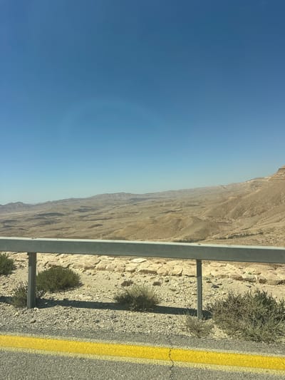 The image depicts a desert landscape under a clear blue sky, likely taken from a vehicle traveling along a road. In the foreground, a grey metal guardrail runs horizontally across the frame, supported by vertical posts. Below the guardrail, a yellow line is painted on the asphalt road, with gravel and sparse desert vegetation at the edge. The middle ground reveals a vast, arid valley with rolling hills and mesas, characterized by muted brown and tan hues. Rocky outcrops and dry, scrubby bushes dot the terrain. The background shows a range of distant mountains, their forms softened by the haze of the desert. The scene is one of a remote, undeveloped area, consistent with the desert environment of Ramat Negev Regional Council, Israel.