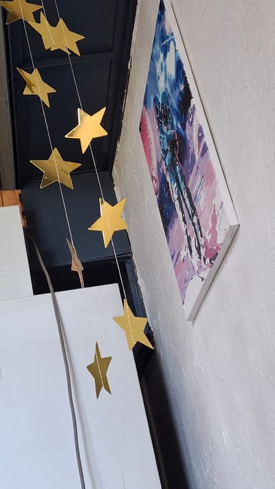 This image captures a domestic interior scene, with no visible indicators of its specific location in Dachi, Kazakhstan. The foreground features a string of gold star ornaments hanging vertically. Below the stars, a large white appliance or surface occupies the left side of the frame. To the right, a wall with a textured finish is visible, adorned with a canvas painting. The painting depicts an astronaut or a figure in a futuristic suit, rendered in a style with bold strokes of blues, purples, and pinks, suggesting a space or sci-fi theme. The painting is hung at an angle, leaning slightly away from the wall. The overall impression is one of simple home decor.