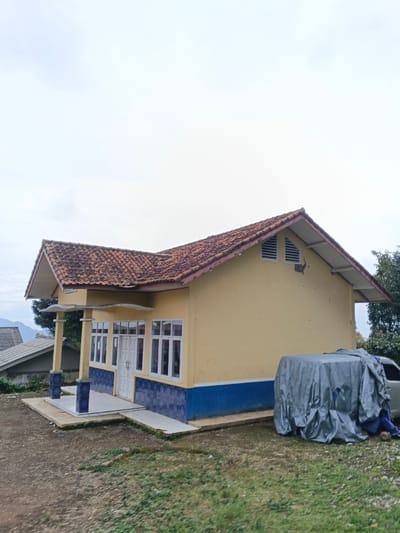 A single-story building with yellow exterior walls and a reddish-brown tiled gable roof is depicted in São Félix do Xingu, Brazil. The building features a prominent blue horizontal stripe along its lower perimeter. The front facade includes a white double door and multiple white-framed windows. A small covered portico, supported by two columns with blue patterned bases, extends over the entrance. The lower sections of the front and left walls are covered with blue patterned tiles. The right gable end of the structure exhibits two horizontal louvered vents near the roof peak and minor damage to the paintwork. To the right of the building, an unidentified vehicle is largely covered by a gray tarp, with a section of blue tarp visible beneath. The immediate foreground consists of uneven dirt ground with scattered patches of green grass. The background reveals distant hills or mountains partially obscured by vegetation, and the roof of another building is visible on the far left. The sky is uniformly overcast. No individuals are present in the scene.