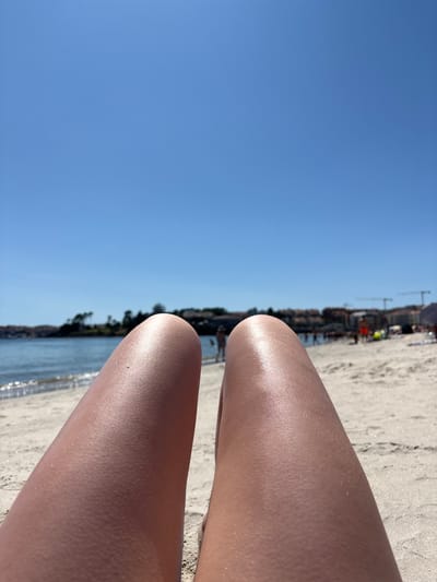 The image depicts a first-person perspective of a person's legs resting on a sandy beach with the ocean and a city skyline in the background. The sky is clear and blue, indicating a sunny day. The water of the ocean is calm, with gentle waves reaching the shore. In the distance, buildings and trees line the coast, suggesting an urban or semi-urban environment. There are a few blurry figures of people visible further down the beach and in the water, engaged in various activities. The beach sand is light in color and appears to be slightly damp near the water's edge. The scene is likely set in Sanxenxo, Spain, known for its beaches and coastal attractions.