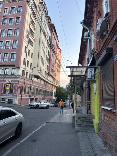 The image depicts a street scene in Sulak, Russia, viewed from a low angle looking upwards. The perspective is from a sidewalk or street level, with buildings dominating the frame. The architecture is varied, featuring brick buildings and a taller, multi-story apartment or office building with many windows. Several air conditioning units are visible mounted on the exteriors of the buildings.
A few people are present in the scene. One person, wearing an orange shirt and blue jeans, is walking away from the camera on the right side of the street. Another person is partially visible further down the street, and a few more figures are in the distance. Several cars are parked or driving on the street, including two silver sedans. Electrical wires are strung across the sky, adding to the urban feel.
The time of day appears to be late afternoon or early evening, with the sunlight casting a warm glow on the buildings. The sky is mostly clear, with a few wispy clouds. The weather seems pleasant.
Visible text includes a sign with "tōga" in white letters on a dark background and a yellow triangular warning sign with a black symbol. Other signs are present but are too small to be legible. The overall impression is a typical urban street in Russia, with a mix of older and newer architecture, daily life unfolding, and the infrastructure of a city.
