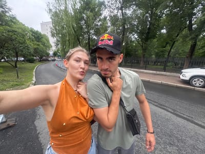 In this image, a young Caucasian couple is posing for a selfie outdoors. The woman, on the left, has blonde hair pulled back and is wearing an orange halter top with gold embellishments and blue jeans. She is puckering her lips as she holds the camera. The man, on the right, has dark hair and is wearing a green t-shirt and a black baseball cap with the Red Bull logo. He has a black crossbody bag slung over his shoulder. They are standing on a paved pathway next to a road. In the background, there are trees, a tall white building, and parked cars. A white SUV is visible on the right side of the frame. The scene appears to be in a park or residential area. The location is identified as Xiangfang District, China.