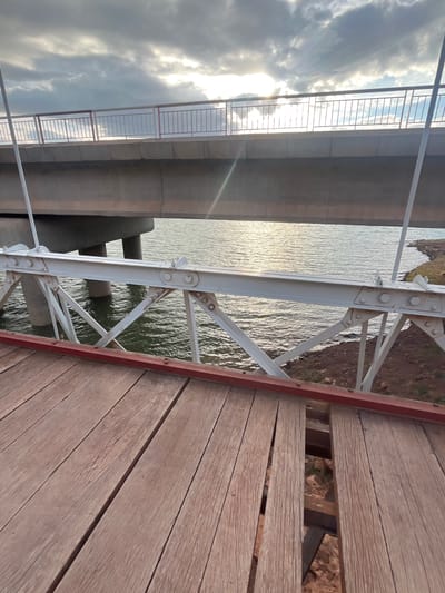 The image presents a view from a wooden bridge, showcasing a steel support structure with white paint, extending towards a body of water with visible reflections. Adjacent to the wooden bridge is a concrete structure with a metal safety barrier, possibly another bridge. The environment includes a glimpse of land on the opposite side of the water. Based on the description of the location, the image is taken in Tabaroucht, Morocco. No people are visible.