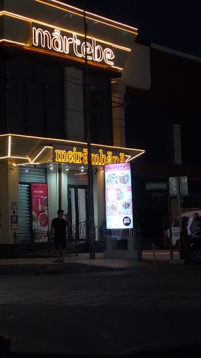 This is a nighttime photo of a restaurant called "Märtebe" in Sarıağaş, Kazakhstan. The building features prominent neon signage, with "Märtebe" written vertically in large, bright yellow letters on the left side of the image. To the right of this, another sign with a pink background displays images of food and the text "meirë mbanëz" and "märtebe by Ana". Below this, a smaller sign advertises "FAST FOOD" with "Coca-Cola + Sprite" logos.
A person is visible standing on the right side of the image, facing away from the camera and towards the building. The street in front of the restaurant is dark, suggesting it is late at night. The lighting from the signs casts a warm glow on the building's facade. The overall scene depicts a commercial establishment illuminated for nighttime operation. One of the signs also contains what appears to be a phone number, "8 702 1".