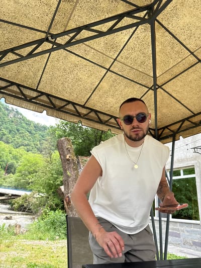 In Vahagni, Armenia, a man stands under a beige, textured canopy with a dark metal frame. He is wearing a white sleeveless shirt, grey pants, sunglasses, and white earbuds. A silver chain with a round pendant is around his neck, and he is wearing rings on his fingers and bracelets on his wrist. He is holding a pink phone in his right hand and looking towards the camera. In the background, a lush, green mountain slopes upwards, with trees and a body of water with a bridge visible. A cut tree trunk is to the left of the man. A building with large windows is behind him on the right.