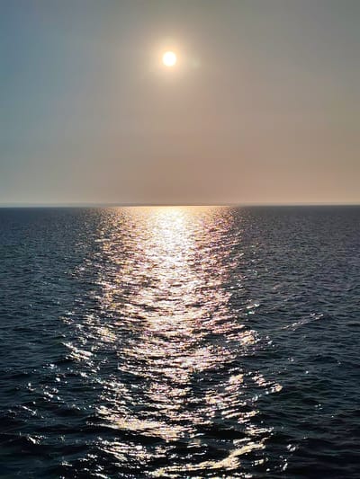 The image depicts a body of water under a bright sun. The sun is positioned in the upper center of the frame, casting a strong reflection on the water's surface, creating a shimmering path of light that stretches towards the viewer. The water appears dark and choppy, with small waves visible. The horizon line is clearly defined, separating the water from a hazy sky that transitions from a light grey to a pale blue. No people, animals, or man-made structures are visible in the image. The scene appears to be a natural landscape, likely an ocean, sea, or large lake. There is no indication of the city of Prislon', Russia, in the image.