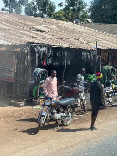 The image depicts an outdoor commercial scene in Tataura, Nigeria. A single-story building with a rusted, corrugated metal roof serves as a tire and inner tube shop, with numerous black rubber items displayed prominently on racks and stacked both inside and outside the open-front structure. The roof's front edge features a series of spikes, and a small vent is visible on the roof surface. A partially obscured white sign with red text on the shop's left side identifies "J. YAYATA COLLEGE & TECHNOLOGY" and advertises "ADMISSION."
Three adult males are present. On the left, a man wearing a light peach-colored tunic and trousers stands beside a dark-colored motorcycle, which is parked on a kickstand on a dirt and gravel patch. In the center, behind the first man, another man in a patterned blue and white shirt stands near a red motorcycle. To the right, a third man, wearing a black sleeveless top, dark shorts, and a red and yellow knitted cap, stands with his back partially to the camera, facing the shop. A silver-colored motorcycle is partially visible behind him. The ground transitions from dirt and gravel near the shop to a paved surface in the immediate foreground. Bright sunlight casts distinct shadows. Green foliage and trees are visible in the background under a clear sky.