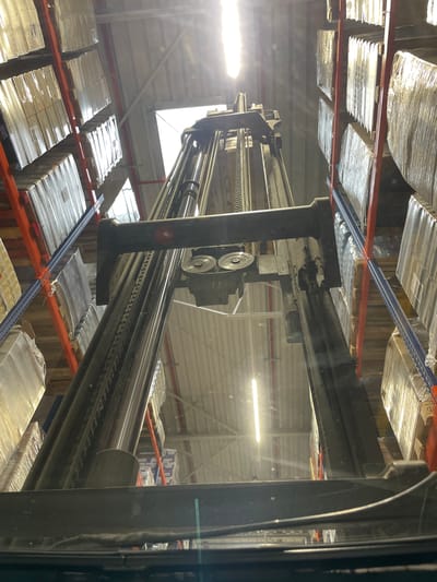The image depicts the interior of a warehouse in Nowa Wieś Kącka, Poland. A forklift is positioned centrally, its mast extending upwards. Tall, multi-tiered shelving units filled with pallets wrapped in plastic are visible on both sides of the forklift. The lighting suggests it might be daytime, with light beams entering from above, possibly through skylights or a large opening. The overall impression is one of industrial storage and logistics. There are no people visible in the image.
