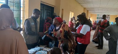 A group of people are gathered inside a building that appears to be a community center or a school in Sabon Gari South, Nigeria. The room has a high ceiling with visible electrical conduits and fluorescent lights. The walls are painted in a light color, and there are windows with metal bars. In the foreground, a man wearing a beige vest with logos is handing an item to a woman in a colorful dress and a red headscarf. There are several packages, possibly containing supplies, laid out on a table or surface in front of them. Behind them, other individuals are standing, some in traditional attire and others in more casual clothing. A woman wearing a red vest over a white shirt and a black baseball cap is also visible, interacting with someone off-camera. Further back, a banner is displayed on the wall, indicating some kind of distribution event, possibly related to children or education, with text that includes "DISTRIBUTION OF..." and "LEARNERS WITH DISABILITY". The overall scene suggests an organized distribution of aid or resources within a community setting.