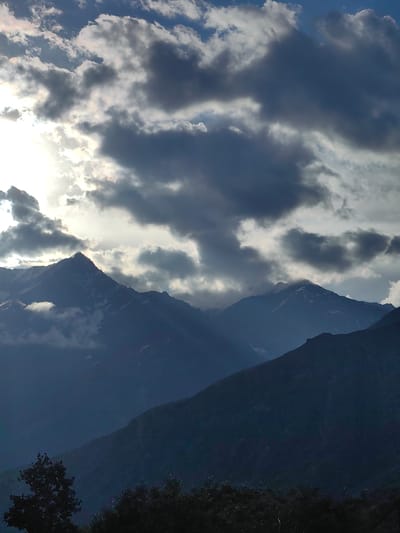 The image captures a majestic mountain range under a dramatic sky filled with dark, heavy clouds interspersed with bright patches of sunlight. The foreground shows a silhouette of dark trees. The mountains, some capped with snow, recede into the distance, their peaks fading into the dusky atmosphere. The lighting suggests either sunrise or sunset, with the sun's rays piercing through the clouds, creating a sense of depth and highlighting the mountain contours. The scene is devoid of human presence and any signs of urban activity, presenting a purely natural landscape. The location is identified as Tiling, India, which is known for its stunning Himalayan vistas, making this a characteristic scene for the region.