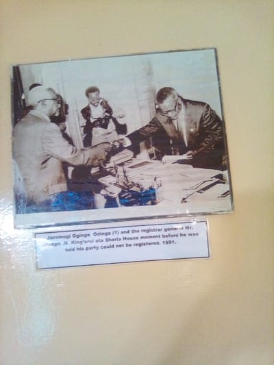 This photographic image, observed in South Sakwa ward, Kenya, displays a historical scene depicting three men in an indoor office environment, identified by the accompanying caption as Sheria House in 1991. In the foreground left, Jaramogi Oginga Odinga, wearing a light-colored suit, spectacles, and a hat, leans forward, his right hand positioned over a desk densely covered with multiple stacks of documents and files. Facing him on the right, Registrar General Mr. Joseph N. King'arui, dressed in a dark suit and spectacles, is also observed leaning over the desk, appearing to examine papers. In the center background, a third man, partially out of focus, is positioned holding what appears to be a camera or recording device. The caption specifies this moment occurred just before Odinga's party was denied registration.