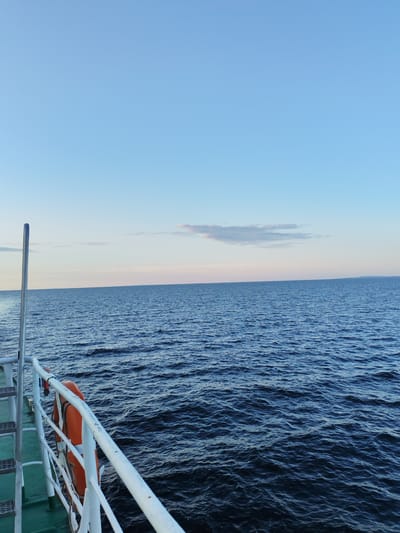 The image shows a view from the deck of a vessel on the water, likely a ferry or a small ship. The sky is clear and a pale blue, with a hint of pink near the horizon, suggesting it is either early morning or late evening. A few wispy clouds are visible. The sea is a deep blue, with gentle waves. In the foreground, part of the ship's railing and a life preserver are visible. The life preserver is orange and appears to be mounted on the railing. The deck of the ship has a green painted surface and white railings, with metal stairs leading down. The location is identified as Kakkarovo, Russia, but this specific information cannot be visually confirmed from the image itself. There are no people visible in the frame.