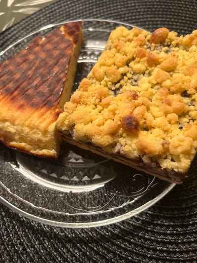 Two slices of cake are presented on a clear, patterned glass plate, which rests on a dark, textured placemat. The slice on the left is a thick, triangular piece with a golden-brown, slightly torched top, revealing a dense, pale yellow interior. The slice on the right is a rectangular piece topped with a generous crumble of golden-yellow pastry. Beneath the crumble, a dark layer, possibly jam or fruit filling, is visible. The background is out of focus, showing hints of a white and green patterned surface. No people are present in the image. The setting appears to be a domestic or café environment, and no specific location in Środa Wielkopolska, Poland, can be identified from the visual information.