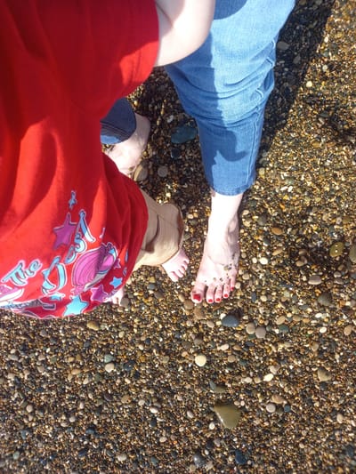 This image is a high-angle shot of two pairs of bare feet on a pebbly beach. The feet are positioned close together, with one larger adult foot on the right and a smaller child's foot partially visible on the left. The adult foot has red nail polish on its toenails. The child's foot appears to be wearing a light-colored sandal. The background consists of many small, smooth pebbles of various colors, including brown, gray, and white. The surface of the pebbles is wet, suggesting the presence of water. The lighting indicates it is daytime, with sunlight casting shadows. The image does not contain any specific landmarks or clues to definitively place it in Gwandra, Abkhazia, or any city.